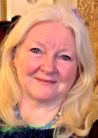 Close-up portrait of a smiling older woman with long blonde hair, blue eyes, and a turquoise beaded necklace, indoors.